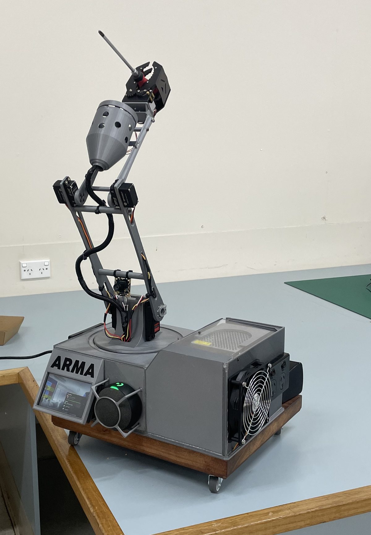 ARMA, a grey 3D-printed robotic arm on a hardwood base, holding a screwdriver in its claw